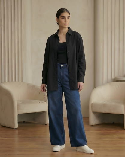 Woman wearing a black shirt, dark jacket, and blue jeans standing in a room with beige walls and furniture.