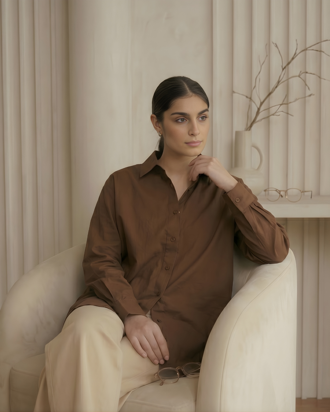 Woman sitting on a white chair wearing a brown shirt and beige pants in a neutral-colored room.