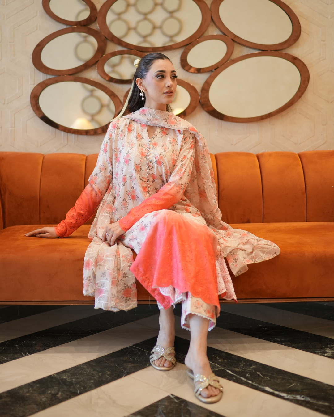 Woman in a floral dress sitting on an orange couch with decorative wall mirrors in the background.